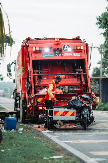 A worker wearing a high-visibility yellow and orange vest is standing at the rear of a large red refuse collection truck, which is positioned on the side of a street. The truck features a white circular logo and various safety stickers, with an open rear compartment revealing a mechanism for collection and unloading waste. The worker appears to be managing waste or equipment, with a black plastic bag placed on a small platform attached to the vehicle. Nearby, a motorbike is parked close to the truck, with a black saddlebag attached to its seat. The environment includes a pavement and a grassy strip, with trees and power lines visible in the background under an overcast sky. This scene illustrates a private waste collection operation, suggesting an alternative to council rubbish disposal, supporting the services provided by Rubbish Removal Westminster for on-site clearance or independent collection in Westminster SW1.