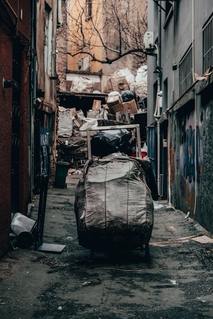 A narrow alleyway in an urban environment filled with overflowing waste and discarded materials. In the foreground, a large, grey, weathered garbage sack is mounted on a metal trolley, positioned centrally on the uneven pavement. The sack appears to contain mixed rubbish, with visible creases and dirt marks on its fabric surface. To the left of the trolley, a small black waste bin is tipped over on the ground, with some identified debris scattered around it. The alley is flanked by tall buildings with graffiti on the lower walls and exposed utility cables running along the facades. In the background, a chaotic pile of cardboard, plastic bags, and other refuse is stacked against a makeshift barrier, creating a cluttered scene indicative of a private waste collection or on-site rubbish clearance. The lighting is muted, suggesting an overcast day, and the environment overall reflects an area in need of waste removal services like those provided by Rubbish Removal Westminster, illustrating a typical example of urban rubbish accumulation in need of professional clearance to support alternative waste handling methods.