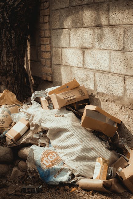 A pile of black rubbish bags, cardboard boxes, and miscellaneous waste materials stacked in front of a red door on a building's exterior wall. The door has graffiti markings, a white sign indicating 'No Money' and 'Keep Clear' in blue, and is framed by a narrow concrete surround. The waste includes torn cardboard packaging and plastic bags, some partially opened, revealing their contents. The scene is set outdoors near a pavement or alleyway, with the rubbish accumulation obstructing part of the wall and door area, illustrating an instance of unmanaged or excess waste that a private rubbish removal service like Rubbish Removal Westminster might handle during on-site clearance or alternative waste management efforts.