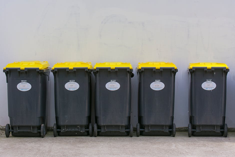 A row of five large wheeled rubbish bins positioned in front of a plain, light-colored wall in an outdoor area, possibly a driveway or communal space. The bins are made of dark grey plastic with textured surfaces and are fitted with bright yellow lids that appear smooth and slightly reflective. Each bin has a white oval label with printed numbers and partially visible text, indicating their identification or waste type. The bins are evenly spaced, with the first and last bins slightly angled inward, and are standing on a concrete or paved surface. The lighting is natural, casting minimal shadows, and the environment suggests they are ready for collection or disposal as part of a waste management process. The setting is neutral and functional, aligning with practices around independent rubbish removal services or private waste handling, and the scene emphasizes the importance of organized waste storage for proper rubbish disposal in compliance with local regulations.